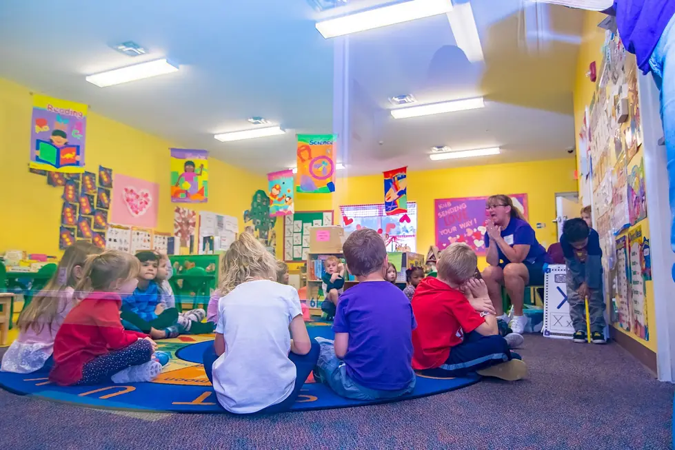 Children seated on the rug for circle time, seen from behind, with a teacher at the edge of the circle